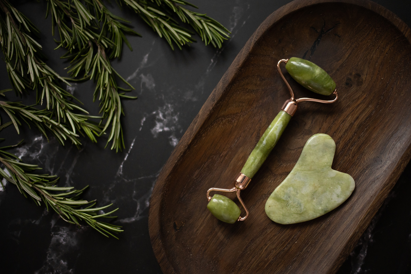 Facial massager with gua sha on timber tray near rosemary